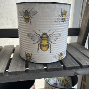 May include: White metal planter with a black rim, featuring a repeating pattern of yellow and black bees. The planter is made from a recycled can.