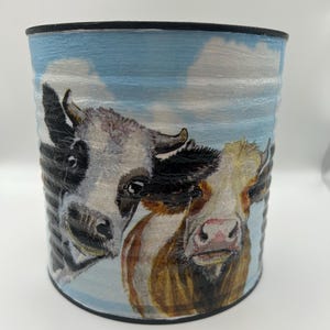 May include: A cylindrical tin can planter with a blue sky and white clouds background. The planter features a painted image of two cows with black and white markings, looking out at the viewer.