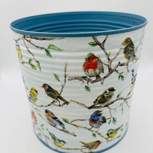 May include: A cylindrical metal planter with a white exterior featuring a detailed pattern of various colourful birds perched on brown branches with green leaves. The interior and the top rim of the planter are painted in a solid matte blue colour.