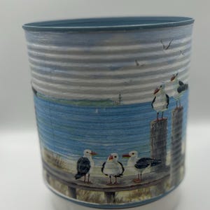 May include: A cylindrical container depicting a seaside scene with seagulls on wooden posts and a beach. The container has a blue rim and base, with a white and blue body.
