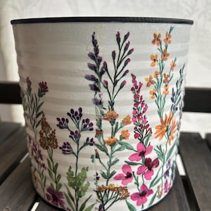 May include: A white, cylindrical plant pot with a black rim, decorated with a colourful floral pattern. The flowers feature purple, pink, orange, and yellow hues, making it a vibrant decorative item for the home or garden.