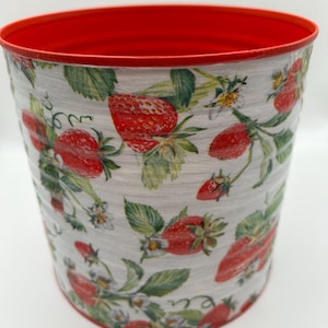May include: A cylindrical container with a red interior and rim. The exterior features a pattern of red strawberries, green leaves, and white blossoms on a light grey background. The container is likely made of metal.
