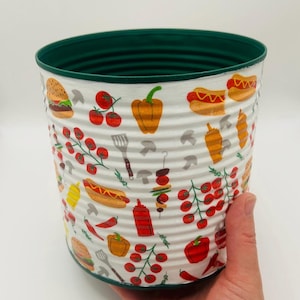 May include: A cylindrical metal container with a dark green rim and interior. The white exterior features a repeating pattern of colourful food illustrations, including hamburgers, hot dogs, red tomatoes on vines, peppers, and grilling utensils.