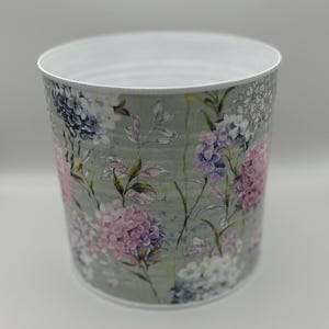 May include: A cylindrical metal container with a white interior and a floral design. The exterior features a light green background with pink, purple, blue, and white hydrangea flowers and green leaves.