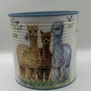 May include: A cylindrical container with a blue rim and a decorative illustration of three alpacas. The alpacas are in shades of brown and blue, standing in a grassy area with floral and bird accents.