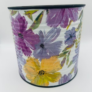 May include: A cylindrical metal container featuring a white background decorated with a painted floral pattern in shades of purple, lavender, and yellow, accented by green leaves and a solid black rim along the top and bottom edges.
