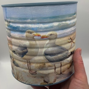 May include: A cylindrical container featuring a painted coastal scene. Two seagulls with orange beaks stand on a sandy beach, with seashells and a blue ocean in the background. The rim is painted light blue.
