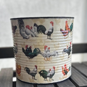 May include: A white metal planter with a black rim, featuring a repeating pattern of roosters and hens. The planter is decorated with a vintage-style design.