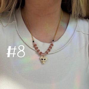 May include: A gold-colored chain necklace with a pendant of a small, off-white dog figurine. The necklace features pink and brown beads. The number "#8" is printed in white on the left side of the image.