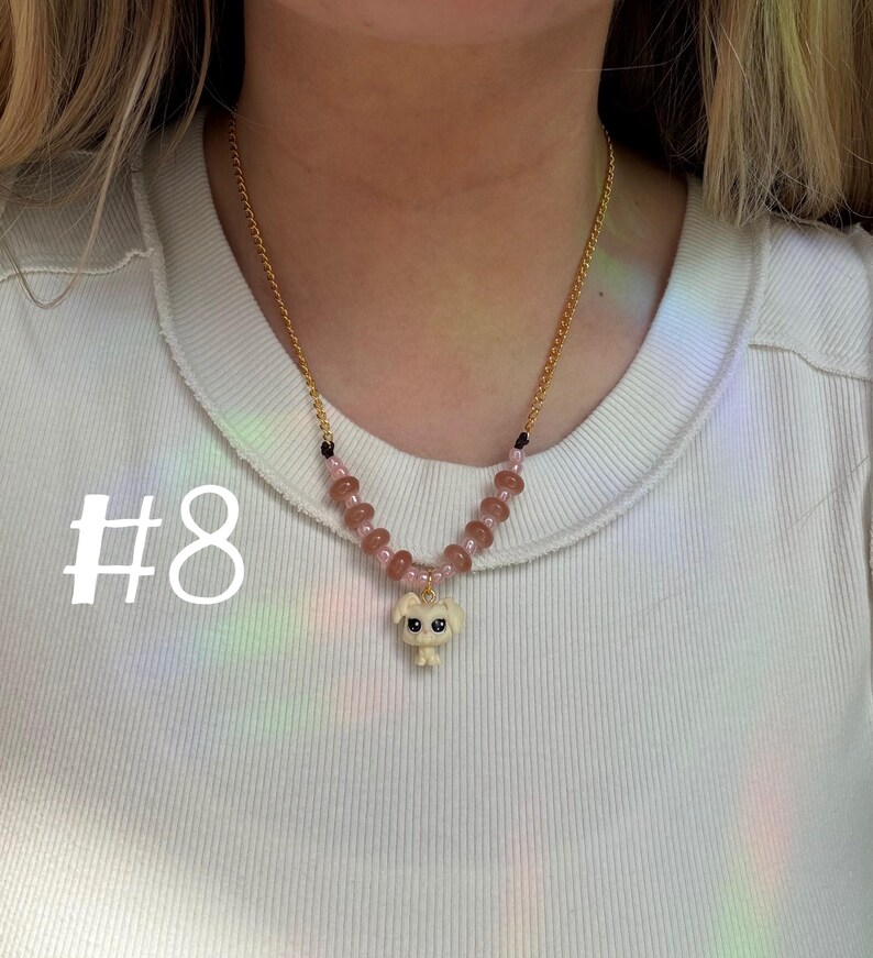 May include: A gold-colored chain necklace with a pendant of a small, off-white dog figurine. The necklace features pink and brown beads. The number "#8" is printed in white on the left side of the image.