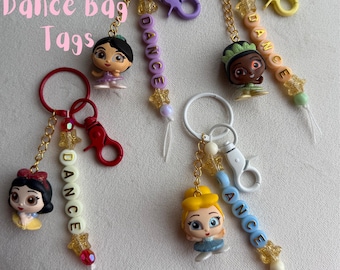 May include: Five princess-themed bag tags with keychains. Each tag features a princess figurine, a beaded word "DANCE", and a decorative charm. The keychains have different colored clasps and rings, including red, purple, yellow, and white.