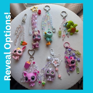 Puede incluir: Una colección de llaveros coloridos con figuras de animales, incluyendo un conejo, un koala y una tortuga. Cada llavero está adornado con cuentas y dijes. El texto "Reveal Options!" se encuentra a la izquierda.