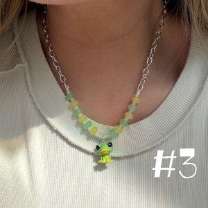 May include: A beaded necklace with a silver chain and a small, green frog pendant. The necklace features yellow and green beads. The number "#3" is visible in the lower right corner.