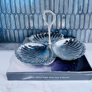 May include: A silver-plated triple shell dish with a central handle, featuring three scalloped compartments. The product is displayed on its original black box, which is clearly labeled with the text: Silver Plated Triple Shell Dish W/Handle.
