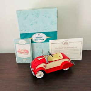 May include: A red and cream Hallmark Kiddie Car Classics 1934 Chrysler Airflow toy car with a black steering wheel, white-walled tires, a matching light blue storage box, a small informational booklet, and a Certificate of Authenticity.