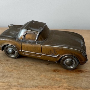 May include: A bronze-colored vintage car model with a hardtop. The car has detailed wheels and a classic design, sitting on a wooden surface. The car's paint has a weathered appearance.