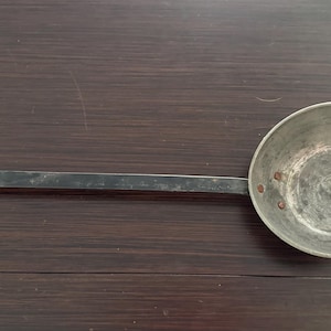 May include: A vintage metal ladle with a long, rectangular handle and a circular bowl. The bowl is riveted to the handle and has a slightly aged appearance. The ladle is likely made of iron or a similar metal.