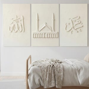 Puede incluir: Tres paneles de arte mural blanquecinos con diseños en relieve. Los paneles presentan caligrafía árabe y una ilustración de una mezquita. La obra de arte se exhibe sobre una cama con un marco de madera y ropa de cama de tonos neutros.