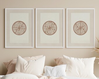 May include: Three framed art prints with a circular design in a muted rose color. The prints are displayed above a bed with white pillows and a striped comforter. The frames are white, and the background is a neutral beige.