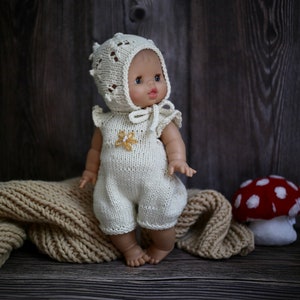 May include: A knitted doll wearing a white crocheted hat and a white knitted jumpsuit with a yellow flower design. The doll is sitting on a brown knitted blanket with a red and white mushroom in the background.