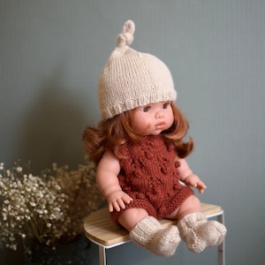 May include: A doll wearing a knitted brown romper with a white knitted hat and matching socks. The doll has red hair and is sitting on a wooden stool.