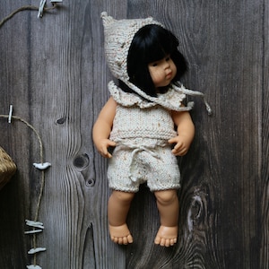 May include: A doll wearing a beige knitted outfit, including a hat, top, and shorts. The outfit has a speckled pattern. The doll has black hair and is posed on a wooden surface. A woven basket and decorative string are in the background.