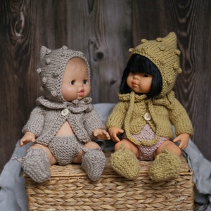 May include: Two knitted dolls wearing matching outfits with hats. The doll on the left is wearing a gray outfit and the doll on the right is wearing a yellow outfit. Both dolls are sitting on a wicker basket.