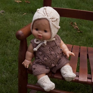May include: A knitted doll wearing a brown and white knitted outfit with a white knitted hat. The doll is sitting on a wooden chair.