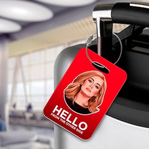 May include: Red luggage tag with a white circle featuring a portrait of Adele. The text "HELLO FROM THE OTHER SIDE" is printed in white on the tag.