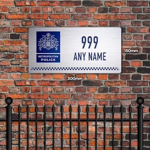 May include: A rectangular metal sign, 30cm x 15cm, with the text "999 ANY NAME" in navy blue. The sign features the Metropolitan Police logo and a checkered border. The sign is mounted on a brick wall.