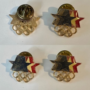 May include: Four gold-colored pins featuring a red, white, and blue star with stripes and the Olympic rings. The back of two pins show the inscription "1984 Los Angeles" and the back of the other two pins show the inscription "84 LA".