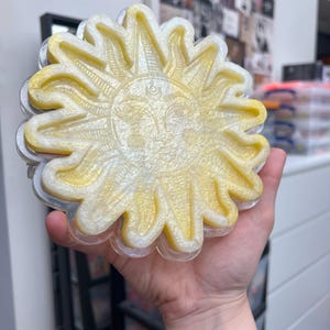 May include: A yellow and white sun-shaped resin coaster with a detailed face and rays. The coaster is made of clear resin with a glittery finish.