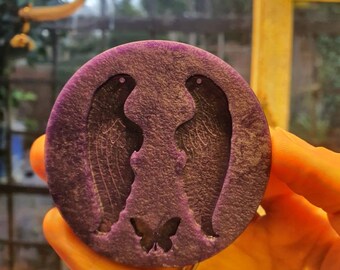 May include: A round, purple, resin coaster with a design of two angel wings and a butterfly in the center.