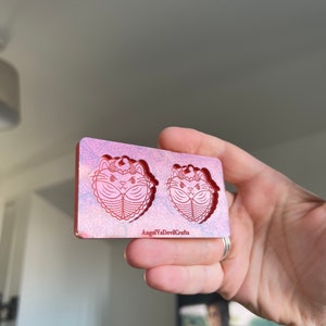 May include: A pink and white resin mold with two cat-shaped cavities. The mold is designed for making earrings and features a detailed cat face with a crown and a heart-shaped body. The mold is labeled "AngelVaDevilCrafts".