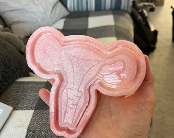 May include: A pink, glittery resin mold in the shape of a uterus. The mold has a detailed interior, including the fallopian tubes and cervix. The mold is held in a hand, with a plaid blanket and pillows in the background.