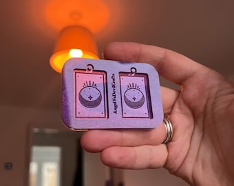 May include: A pair of purple and white resin earrings with a moon and star design. The earrings are rectangular and have a hole at the top for hanging. The text "Angel Wallflower Crafts" is printed on the back of the earrings.