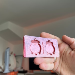 May include: A pink and white resin mould with two cauldron shapes. The mould is for making earrings or other small crafts. The text "Angel & Devil Crafts" is printed on the mould.