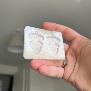 Puede incluir: Un molde de silicona transparente con dos impresiones de un pequeño diseño abstracto detallado. El molde está diseñado para hacer aretes.