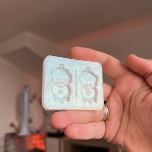 May include: A clear silicone mould with two impressions of a small, round, detailed design. The mould is for making earrings. The text "WeLoveDecals" is printed on the mould.