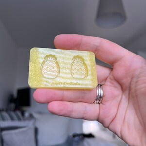 May include: A yellow silicone mold with two pineapple shapes. The mold is in a hand and has the text "AngelVaDevilCrafts" on the bottom.