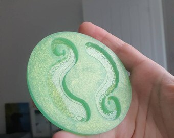 May include: A round, green, translucent resin coaster with a raised design of two octopus tentacles.