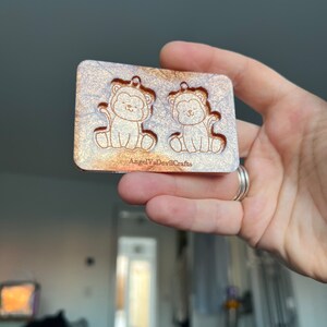 May include: A gold and white resin mold with two small cat designs. The mold is for making earrings or other small crafts. The mold is labeled "AngelVsCrafts".