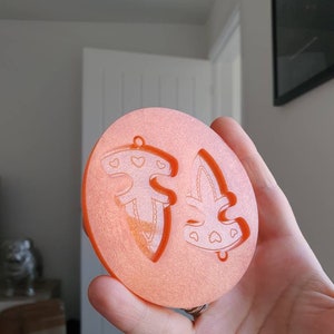 Puede incluir: Un molde de silicona redondo de color naranja con dos formas de árbol de Navidad. Los árboles tienen un diseño de corazón y un agujero en la parte superior para colgar.