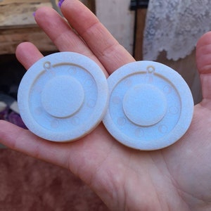 May include: Two white round resin moulds with a raised circle design and a small hole at the top. The moulds are for making moon phase jewellery.
