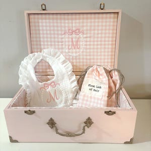 May include: A pink wooden keepsake box with a gingham lining. The box contains a white bib with a pink bow and the letter "M" embroidered on it, and a pink gingham bag with the text "First Lock of Hair" embroidered on it.