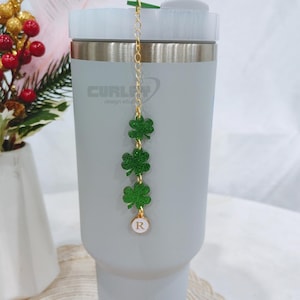 Può includere: Un bicchiere in acciaio inossidabile grigio con una catena dorata e quattro ciondoli a forma di trifoglio verde con glitter. Il ciondolo in fondo alla catena ha un cerchio bianco con la lettera "R" in oro.