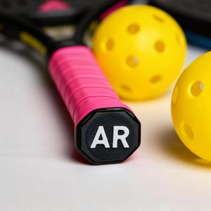 以下が含まれることがあります： ピックルボールの備品のクローズアップ。 ピンクと黒のピックルボールパドルのハンドルで、端に「AR」の文字が入っています。 穴の開いた2つの黄色のピックルボールが背景にあります。
