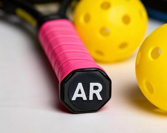 May include: Close-up of pickleball equipment. A pink and black pickleball paddle handle with the letters "AR" on the end is in the foreground. Two yellow pickleballs with holes are in the background.
