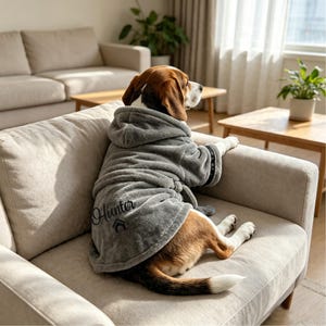 Op de afbeelding: Een beagle-hond draagt een zachte, grijze pluche badjas met capuchon. De badjas heeft lange mouwen, een ceintuur en de naam Hunter geborduurd in zwarte schrijfletters boven een klein zwart huisicoontje op de achterkant van het kledingstuk.