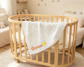 May include: An oval-shaped wooden crib with vertical slats and wheels, featuring a white muslin blanket draped over the side. The blanket is embroidered with the name Ronan in orange cursive script next to a small, yellow, smiling star icon.